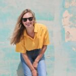 Casual young woman smiling in sunglasses, posing against a vintage colorful wall.